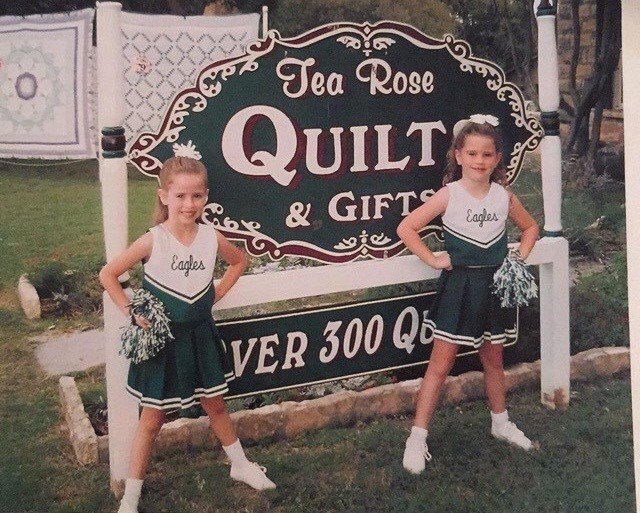 Savannah Sikes and her sister as young Heritage School cheerleaders in front of the Tea Rose Quilts sign in Fredericksburg
